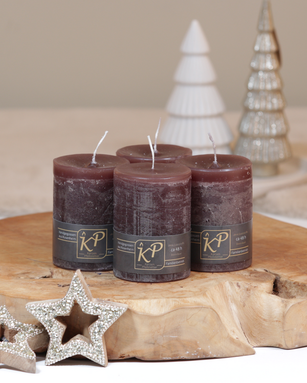 Vier Tischkerzen in dunkelbraun dekorativ auf einem Holztisch mit Weihnachtsdeko im Hintergrund.