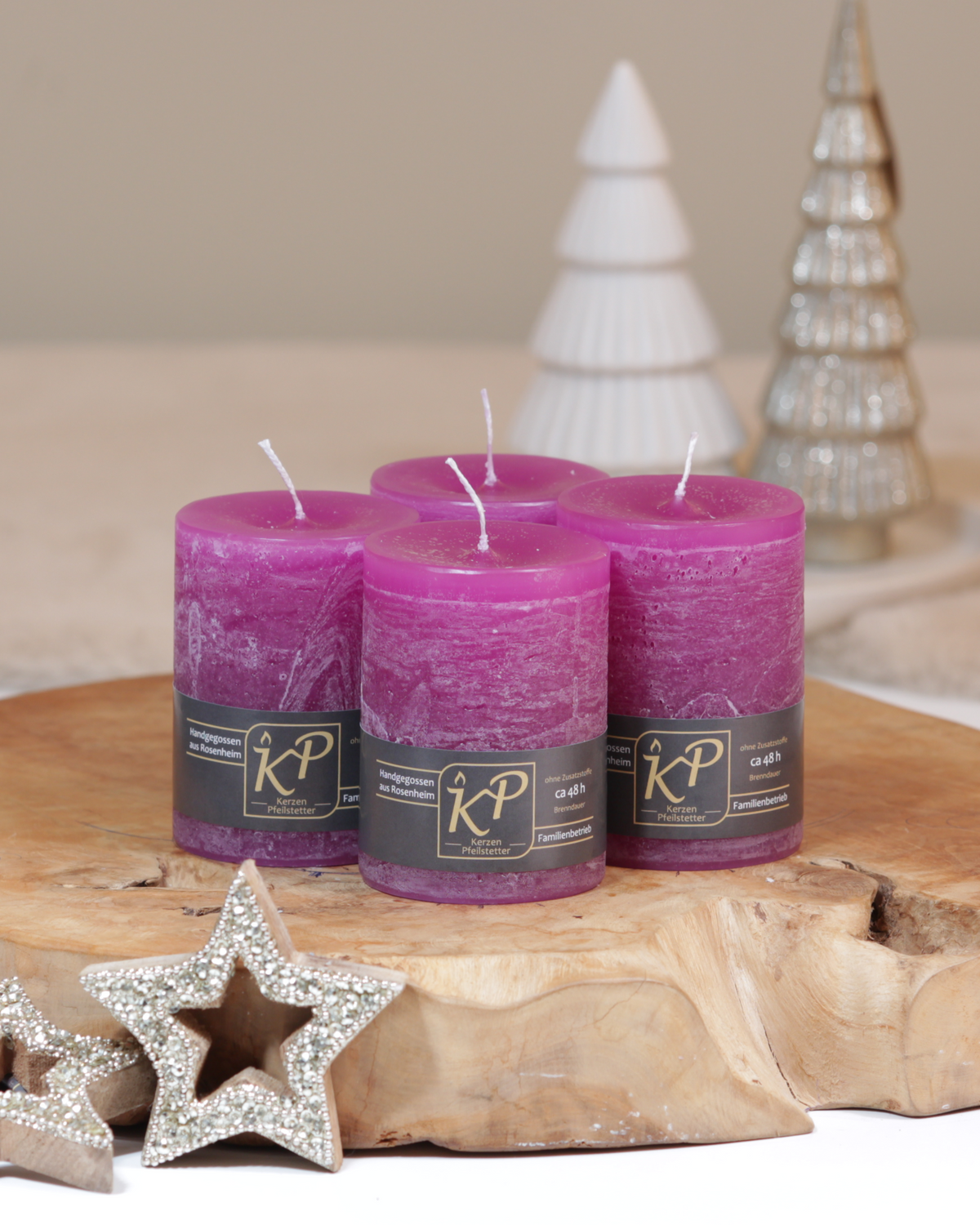 Vier Tischkerzen in magenta dekorativ auf einem Holztisch mit Weihnachtsdeko im Hintergrund.